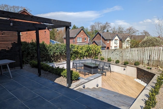 Sleeper walls were replaced with block walls finished in a smooth render, and sandstone paving was replaced with modern porcelain paving. A pergola offers a sheltered patio area and a water feature adds a tranquil backing track. Drainage was installed to improve ground conditions.
